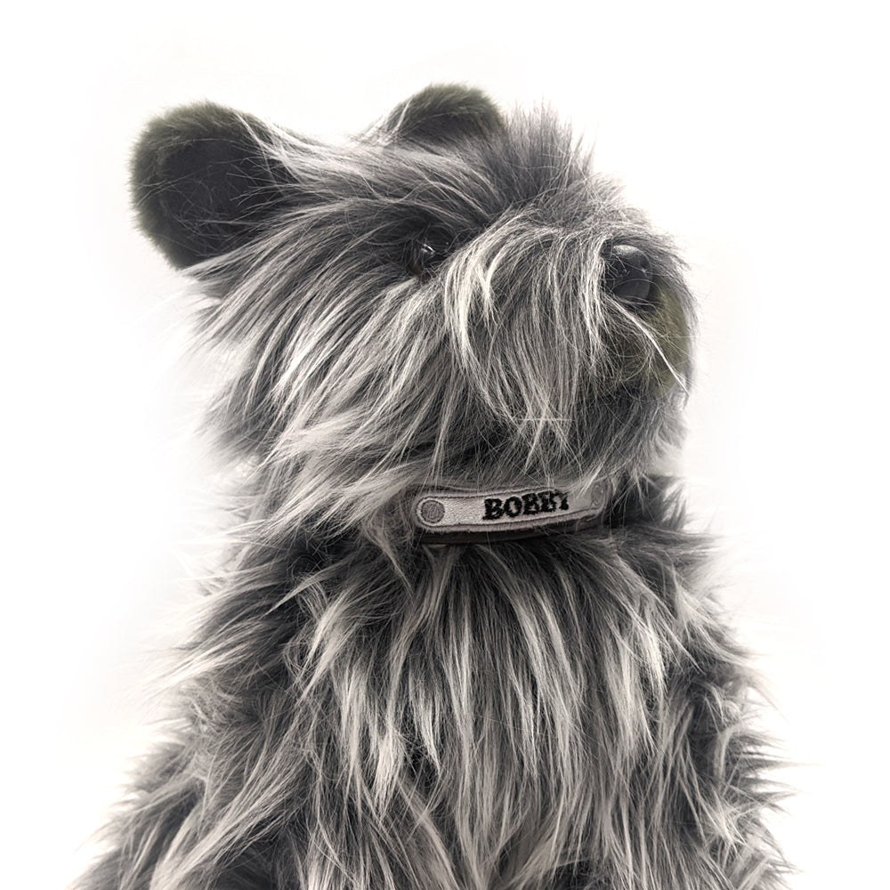 A soft toy of a Skye terrier wearing a collar with the name "Bobby" embroidered on it.