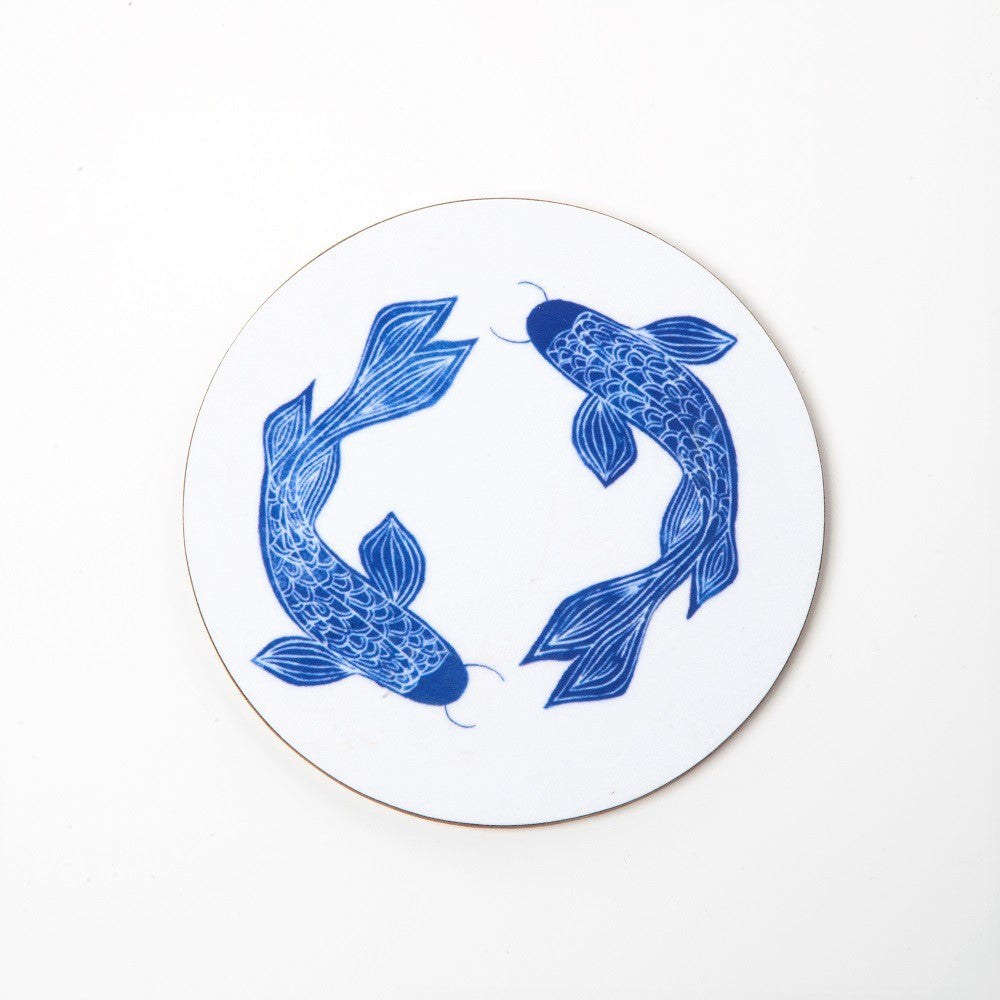 A white round cork backed coaster with illustrations of two blue fish in a Ming style. The fish are in a symmetrical design from above around the coaster.