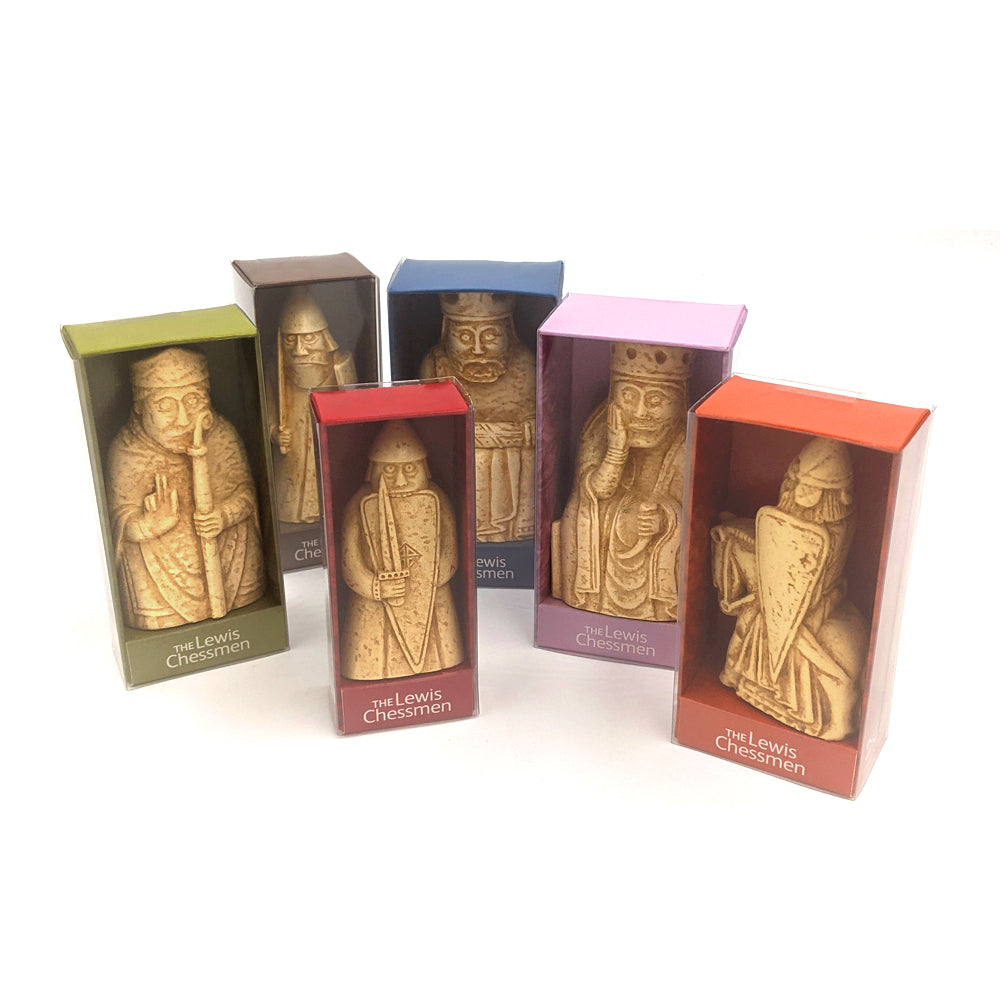 Six colourful cardboard boxes with cellophane windows against a plain white background. Inside the boxes are resin replica Lewis chess pieces. From left to right: A bishop, a warder, a berserker, a king, a queen, and a knight.