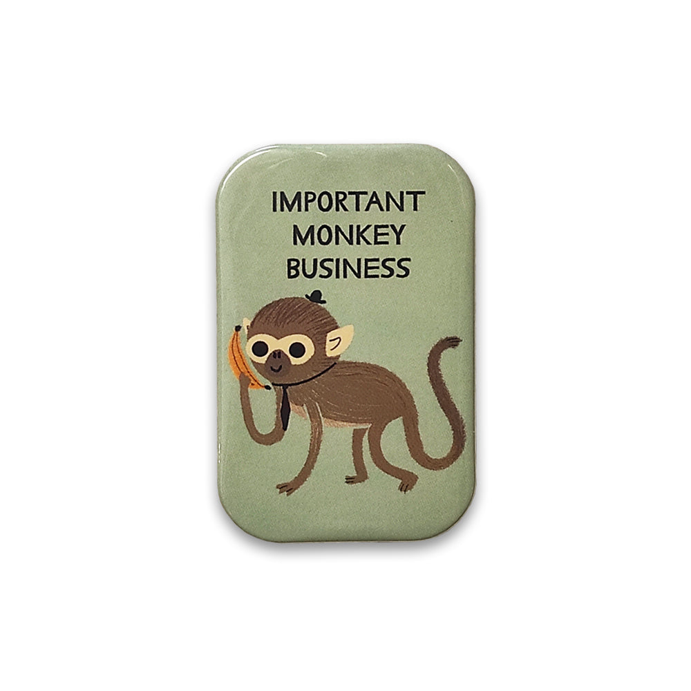 A photograph of a pale green rectangular magnet with rounded corners against a plain white background. The magnet is printed with an illustration of a monkey wearing a hat and tie, and is holding a banana like a phone. The words "important monkey business" are printed above.