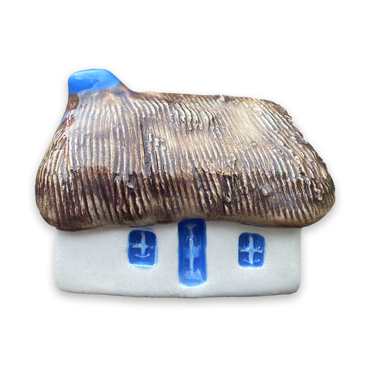 A small white ceramic cottage with a brown painted thatch style roof and a blue door and windows against a plain white background.
