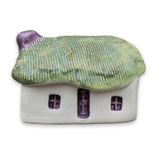 A small white ceramic cottage with a green painted thatch style roof and a purple door and windows against a plain white background.
