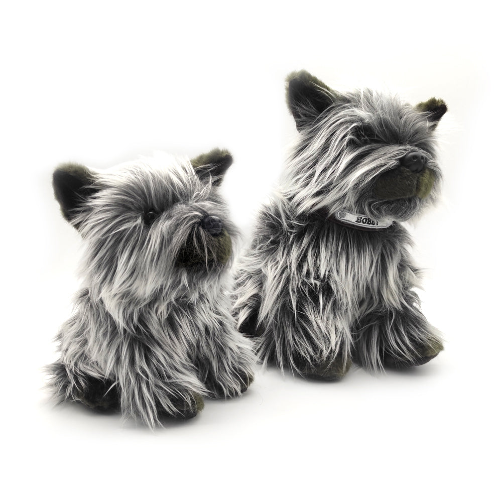 Two soft toy Skye terriers with long grey and black fur against a plain white background. To the left is a smaller dog and on the right, a larger one with a silver collar embroidered with the name "Bobby".