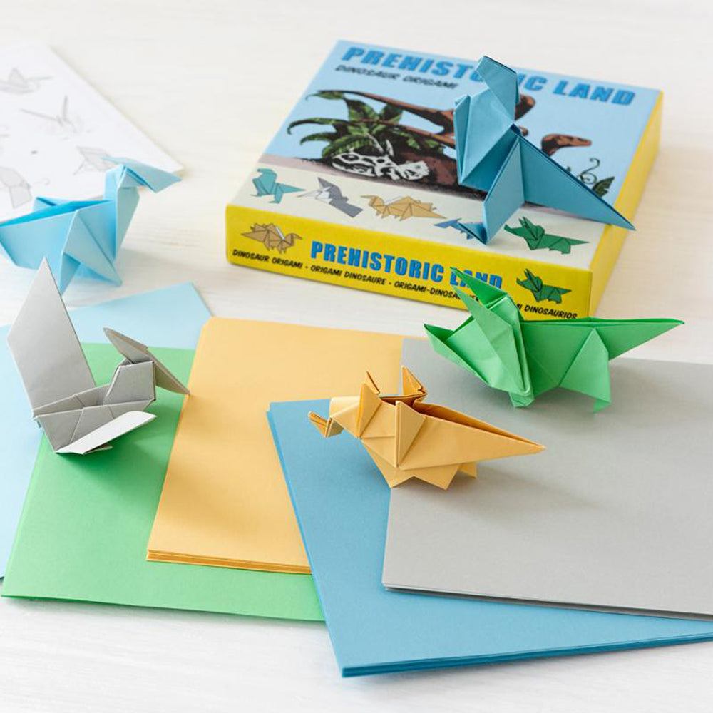 A box and some square sheets of colourful paper on a table. On top of the box and the paper are a selection of small origami dinosaurs