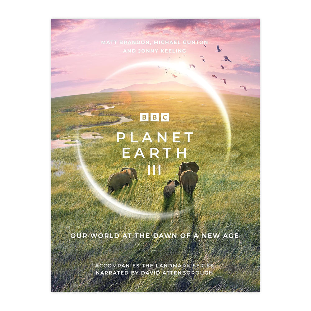 Book cover with a photograph of three elephants walking away from the camera in long grass underneath a lilac sky with birds flying overhead.in the centre a round reflection of light imitating a camera lens with the BBC logo and the words "Planet Earth III" in the centre.