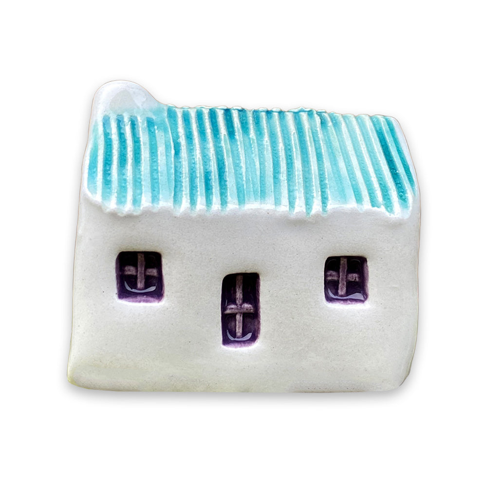 A small white ceramic cottage with a blue painted roof and a purple door and windows against a plain white background.
