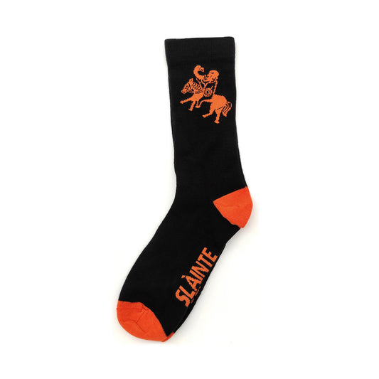 A black sock against a plain white background with a orange toes and heel with an orange image from a carving of a pictish man riding a horse whilst drinking from a horn. The sold of  the socks have the phrase 'Slainte Mhath' also in orange.