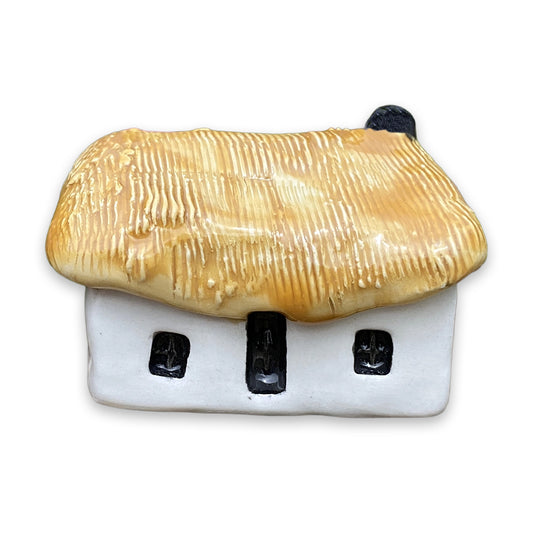 A small white ceramic cottage with a yellow painted thatch style roof and a black door and windows against a plain white background.