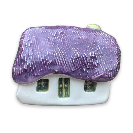 A small white ceramic cottage with a purple painted thatch style roof and a green door and windows against a plain white background.