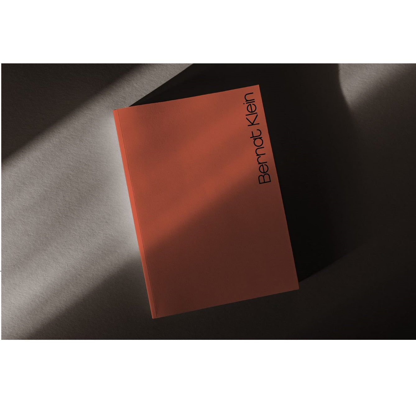 A plain peach coloured book sitting at an angle in a harsh shadow with a bright beam of light passing across it's top half. The top right corner of the book has the words Bernat Klein written vertically along the outer edge.