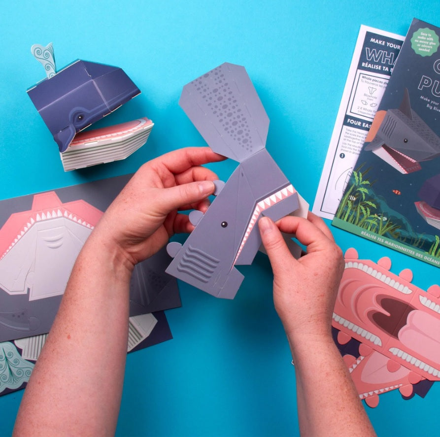 A pair of hands folding and assembling a paper shark hand puppet against a bright blue background.