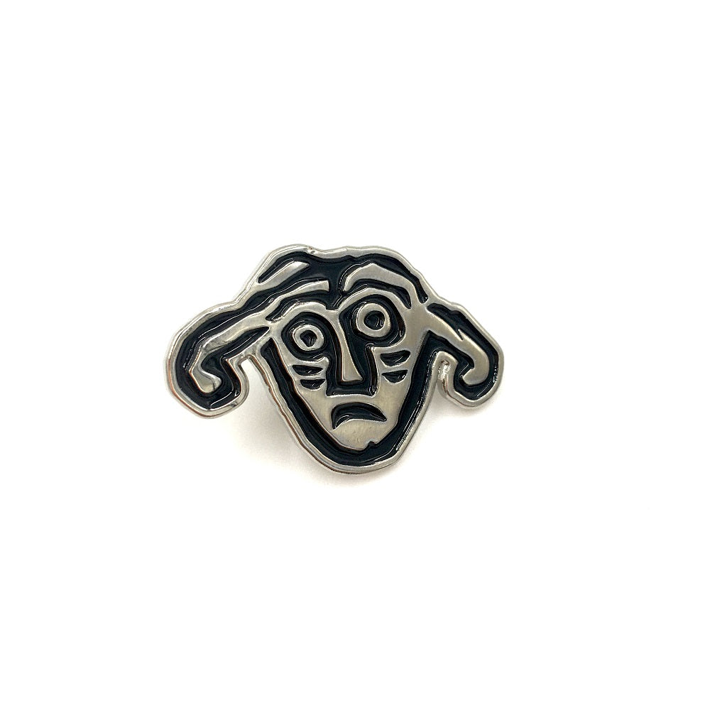 A pin badge with a face from a detail of a brooch from the Galloway hoard. A face with a worried look and two large droopy ears.