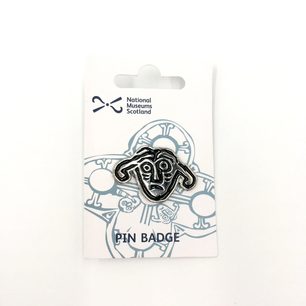 A pin badge with a face from a detail of a brooch from the Galloway hoard. A face with a worried look and two large droopy ears against a white backing card.