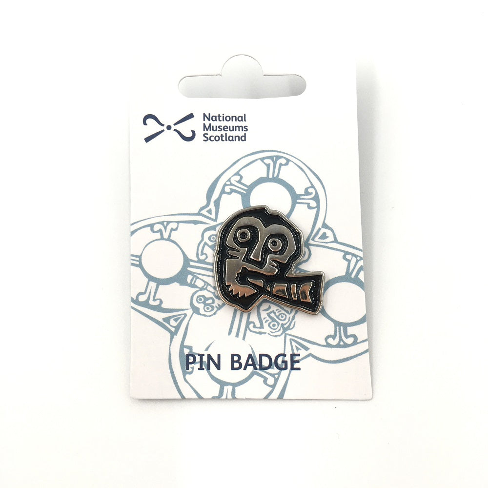 A pin badge with a face from a detail of a brooch from the Galloway hoard. A face playing a horn against a white backing card.