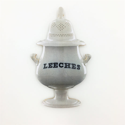 A wooden cut out magnet of a white porcelain jar with the word "Leeches" in black letters across the body of the jar. The jar has two handles on either side and an elaborate lid perforated with holes.