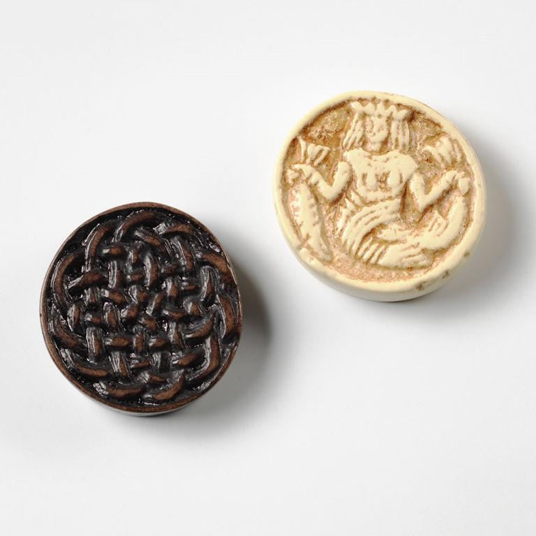 Two counters from the set. One in black with a traditional knot work design, the other depicts a mermaid in a crown holding a fish by the tail in each hand.