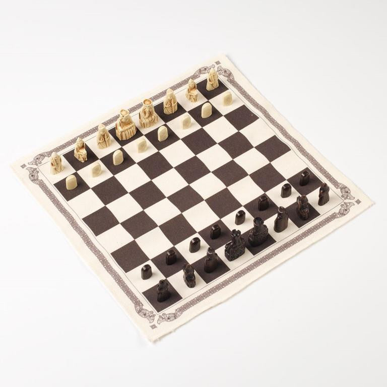 A square cloth chess board border by Norse knot work with black and white chess pieces depicting the Lewis chess pieces.