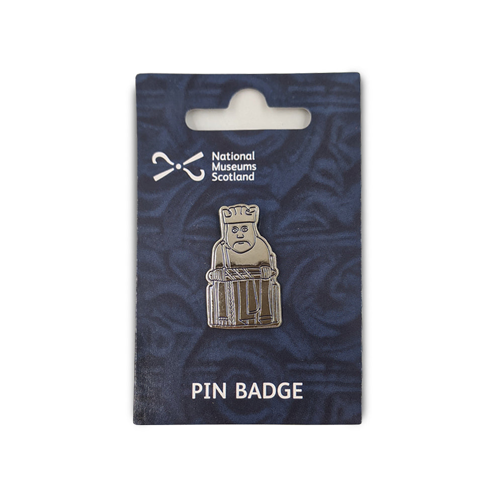 A metal pin badge etched with a line illustration of a king Lewis chess piece. The pin badge is on a navy blue backing card  with a faint knot work design, the National Museums Scotland in the top left and the words "Pin Badge" across the bottom edge in white.